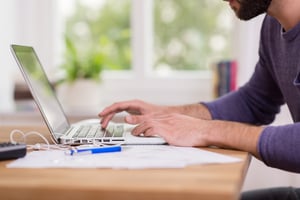 mans hands typing on laptop with home setting in background