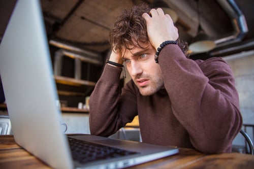 Stressful upset desperate handsome curly man in brown sweatshirt working using laptop and having headache